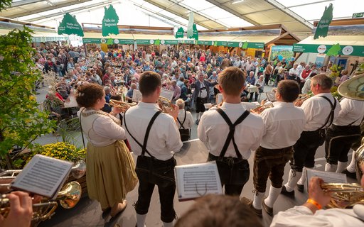 Innenaufnahme einer Messehalle. Musiker mit Blechinstrumenten in Lederhosen und Tracht stehen auf der Bühne, im Hintergrund sind ein großes Publikum und zahlreiche Messestände zu sehen. 