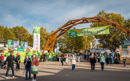 Holztor mit grünem Banner: „Herzlich Willkommen auf dem 101. LWH“. Menschen gehen ein und aus. Fahnen, Banner und Bäume im Hintergrund. 