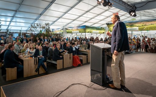 Ein Mann spricht an Rednerpult auf Bühne vor großem Publikum an Tischen in Messehalle. Im Hintergrund weitere Personen und Messestände. 