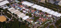 Luftaufnahme des Cannstatter Wasens mit zahlreichen weißen Zelten und bunten Ständen. Viele Menschen bewegen sich über das Gelände, das von Bäumen und Straßen umgeben ist.