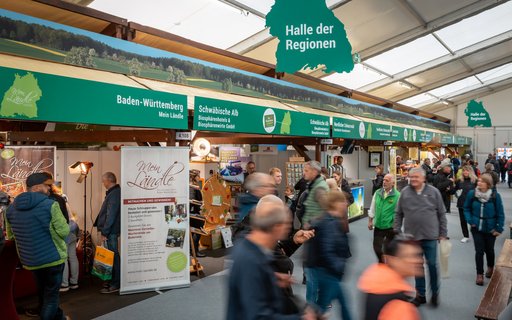 Innenaufnahme einer Messehalle mit Ständen unter dem Schild „Halle der Regionen“ und einer Umrisskarte von Baden-Württemberg. Die Stände tragen Regionalnamen wie „Schwäbische Alb“. Personen gehen hindurch. 