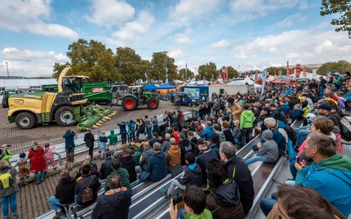 Menschen auf Tribüne schauen Vorführung mit Traktoren und landwirtschaftlichen Geräten zu. Im Hintergrund Zelte, Banner und Bäume bei teils bewölktem Himmel.