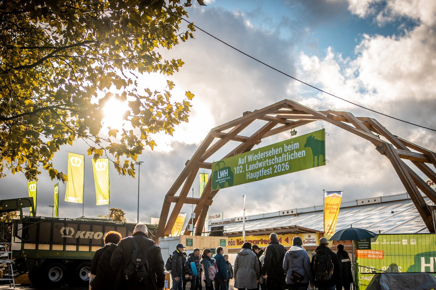 Eingangsportal mit Himmel Gruppe von Menschen unter einem Holzbogen mit grünem Banner: „Auf Wiedersehen beim 102. Landwirtschaftlichen Hauptfest 2026“. Im Hintergrund sind Fahnen, Banner und ein großes Fahrzeug bei teils sonnigem Wetter.