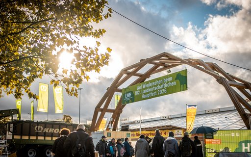 Gruppe von Menschen unter Holzbogen mit Banner: „Auf Wiedersehen beim 102. Landwirtschaftlichen Hauptfest 2026“. Im Hintergrund Fahnen, Banner und ein großes Fahrzeug bei teils bewölktem Himmel.