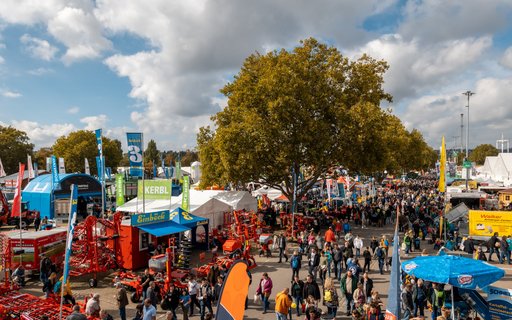 Gut besuchte Messe mit Menschen zwischen Ständen und Ausstellungen. Im Hintergrund Bäume, teils bewölkter Himmel. Bunte Banner, Fahnen und Landmaschinen sind zu sehen.