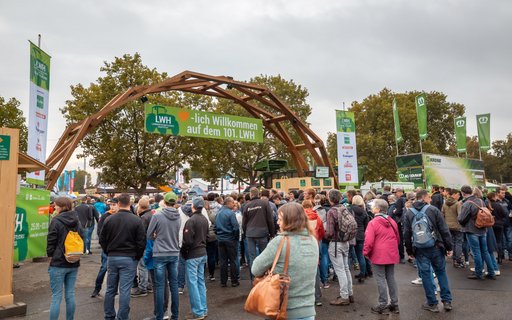 Menschenmenge am Eingang mit Holztor und grünem Banner: „Herzlich Willkommen auf dem 101. LWH“. Umgeben von Fahnen, Bannern und Bäumen im Hintergrund.