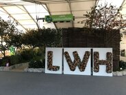 Ausstellung mit drei großen weißen Tafeln, auf denen die Buchstaben ‚LWH‘ aus natürlichen Materialien wie Blättern und Blumen gestaltet sind. Darüber ein grünes Schild mit der Aufschrift ‚LWH Jubiläumsfläche‘. Die Szenerie ist von Pflanzen und Bäumen umgeben und befindet sich im Inneren einer Messehalle.