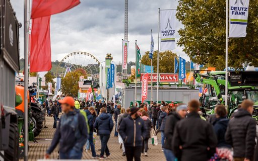Viel besuchtes Messegelände mit Fahnen, Bannern und Ständen für Landmaschinen. Im Hintergrund ein Riesenrad, herbstliche Bäume und bewölkter Himmel. 