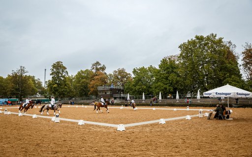 Reitstadion mit mehreren Reiterinnen und Reitern, die Dressurübungen auf ihren Pferden ausführen. Arena ist mit weißen Begrenzungen versehen. Im Hintergrund sind Bäume, Gebäude und Zelte für Zuschauer. Dort sitzen Personen. 