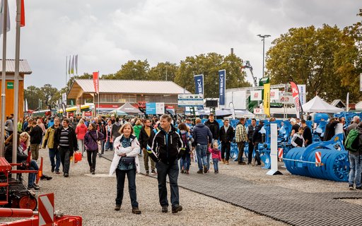 Außengelände einer Messe mit zahlreichen Personen, die zwischen Ständen, Zelten und Ausstellungsflächen umhergehen. Im Hintergrund mehrere Banner, Fahnen sowie landwirtschaftliche Maschinen und der Himmel ist bewölkt. 