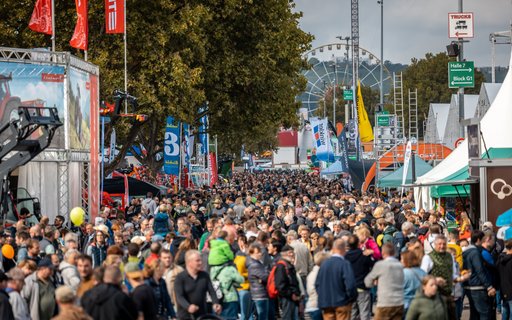 Belebtes Messegelände mit Menschen zwischen Ständen und Zelten. Im Hintergrund Riesenrad, Fahrgeschäfte sowie Fahnen und Banner mit Markennamen.