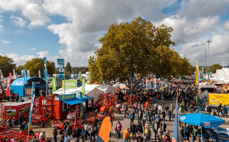 Außenaufnahme einer Messe mit vielen Personen zwischen Ständen und Ausstellungsflächen. Im Hintergrund sind große Bäume und bewölkter Himmel. Zu sehen sind bunte Banner, Fahnen und landwirtschaftliche Maschinen.  