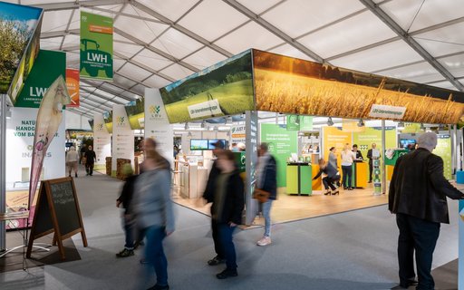 Innenaufnahme einer Messehalle mit Ständen mit den Aufschriften „LWH“, „LBV“ und „LBV-Unternehmensgruppe“. Personen gehen durch die Halle. Über den Ständen hängen große Banner mit Abbildungen von Feldern und regionalen Landschaften.