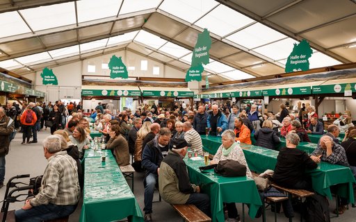 Messehalle mit vielen Menschen an Tischen mit grünen Tischdecken. Über ihnen hängen grüne Schilder in Form von Baden-Württemberg mit der Aufschrift „Halle der Regionen“. Im Hintergrund sind Messestände. 