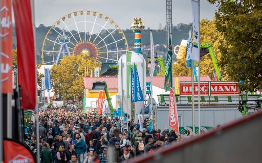 Belebtes Messegelände mit Menschen zwischen Ständen und Zelten. Im Hintergrund Riesenrad, Fahrgeschäfte sowie Fahnen und Banner mit Markennamen.