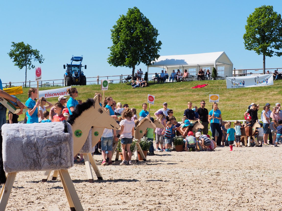 Holzpferde auf einem Reitplatz unter blauem Himmel, Kinder spielen mit den Holzpferden im Hintergrund. 